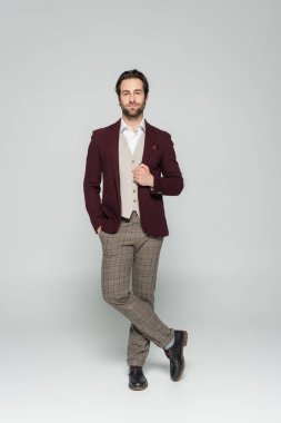 full length of bearded showman in vest and red blazer standing with hand in pocket on grey   clipart