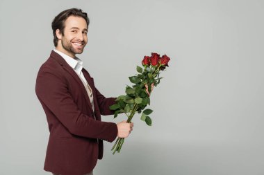 Ceketli neşeli şovmen kırmızı güller tutuyor ve gri renkli kameraya bakıyor. 