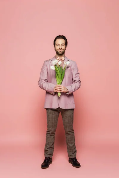 Full length of stylish host of event holding bouquet of tulips on pink background