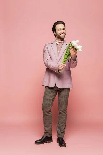 Full length of cheerful host of event holding bouquet of tulips on pink background