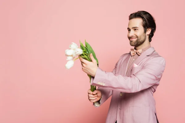 Positive host of event in jacket holding bouquet of tulips and looking away on pink background