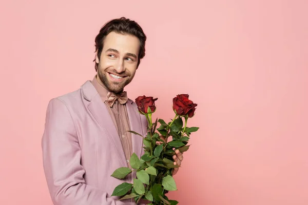 Cheerful host of event holding red roses and looking away isolated on pink