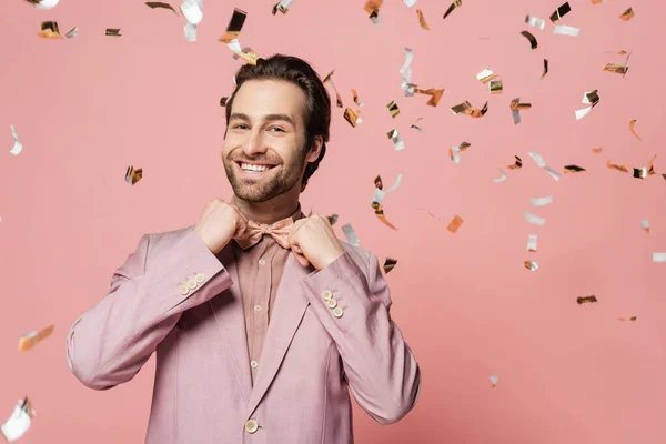 Smiling host of event in jacket adjusting bow tie under confetti on pink background