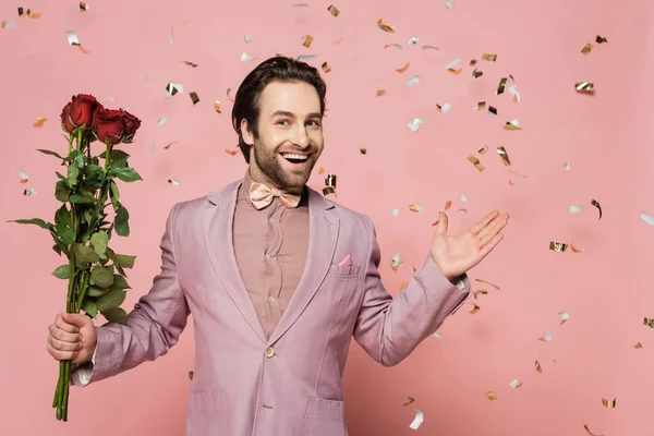 Excited host of event holding roses and pointing with hand under confetti on pink background