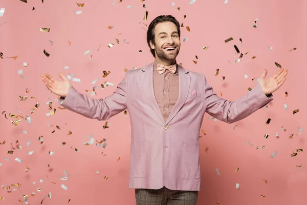 Cheerful host of event pointing at falling confetti and looking at camera on pink background