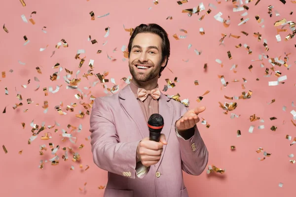 Smiling host of event holding microphone and pointing at camera under confetti on pink background