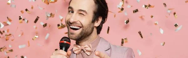 Stylish host of event holding microphone and winking at camera under confetti on pink background, banner