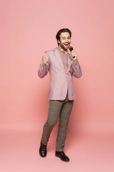 Full length of trendy host of event showing yes gesture and holding microphone on pink background