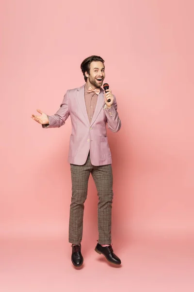 Full length of trendy host of event holding microphone while jumping on pink background