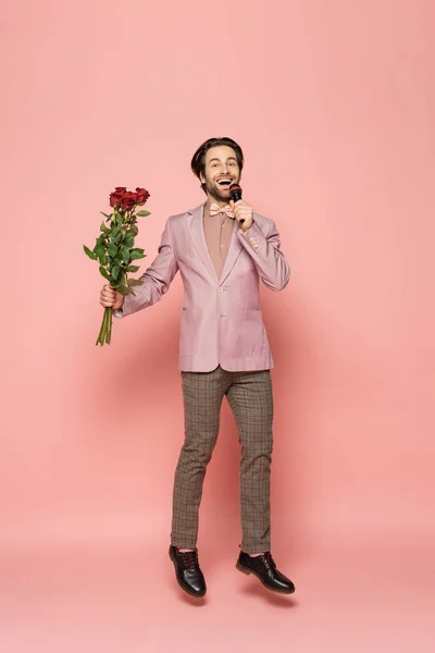 Positive host of event holding flowers and microphone while jumping on pink background