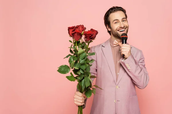 Cheerful host of event holding microphone and bouquet of roses isolated on pink