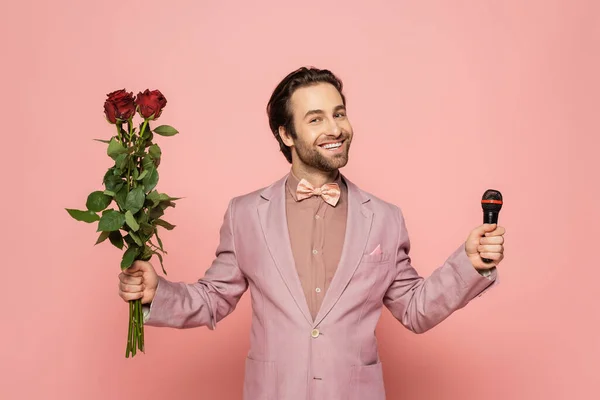Stylish host of event in jacket and bow tie holding microphone and flowers on pink background
