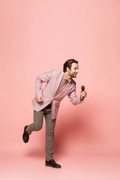 Full length of cheerful and stylish host of event holding microphone on pink background