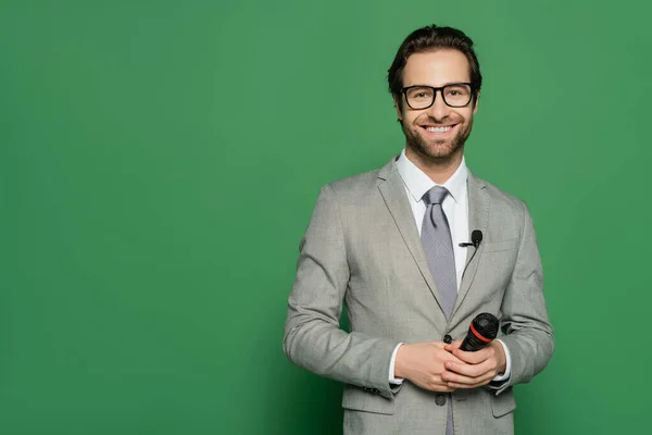 stock image cheerful news anchor in suit and eyeglasses holding microphone on green background 