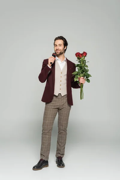 full length of cheerful host of event in blazer holding red roses and talking in microphone on grey