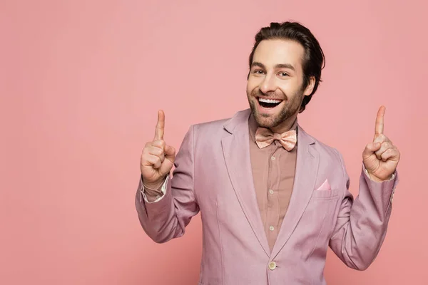 stock image cheerful and young showman in suit with bow tie pointing up with fingers isolated on pink 