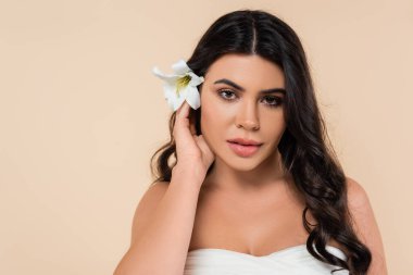 Portrait of brunette woman with lily flower in hair looking at camera isolated on beige  clipart