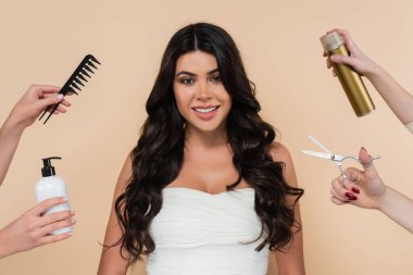 Smiling woman with wavy hair looking at camera near hands with hairdressing tools isolated on beige  clipart