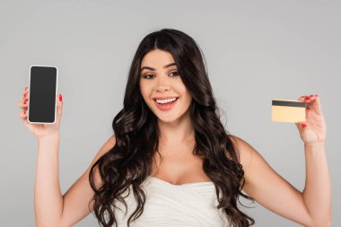 Uzun saçlı neşeli esmer kadın kredi kartı ve akıllı telefon gösteriyor. Beyaz ekran gri üzerinde izole edilmiş.