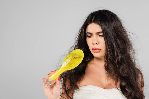 sad brunette woman with tangled hair looking at hair brush isolated on grey