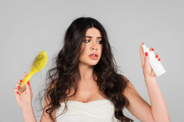 displeased woman with damaged hair holding treatment spray and hair brush isolated on grey