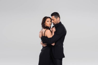 Siyah elbiseli esmer kız arkadaşına sarılan sakallı bir adam, gri alanda izole edilmiş. 