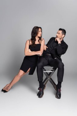 Uzun boylu, sakallı bir adam sandalyeye oturmuş siyah elbiseli esmer bir kadına ve gri topuklu bir kadına bakıyor. 