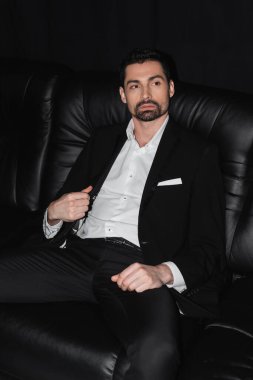 Elegant man in suit sitting on leather couch isolated on black  clipart