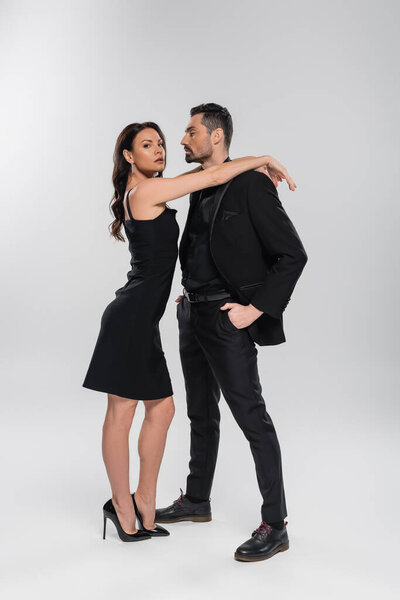 full length of sexy woman in black dress leaning on man posing with hands in pockets on grey