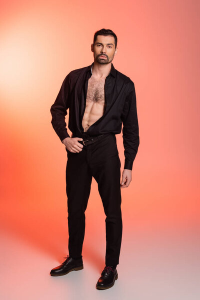 full length of sexy and bearded man with hairy chest standing in black outfit on pink 