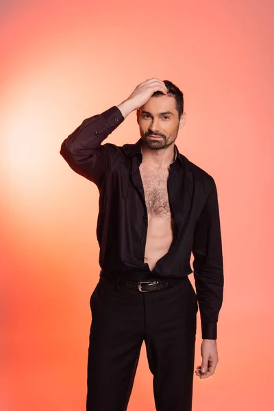 bearded man with hairy chest standing in black outfit and adjusting hair on pink