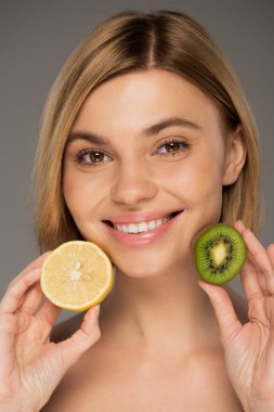Gri renkte kivi meyvesi ve limon tutan çıplak omuzlu neşeli genç bir kadın. 