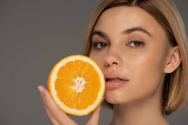 portrait of young blonde woman holding juicy orange half isolated on grey  clipart