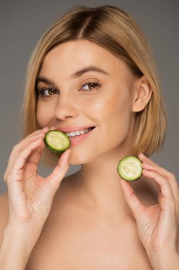 Dilimlenmiş salatalıklar ve gri üzerinde izole edilmiş taze salatalık tutan neşeli kadın portresi 