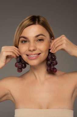 Çıplak omuzlu, kulaklarının yakınında tatlı üzümler tutan mutlu genç bir kadın. Gri üzerinde izole edilmiş. 