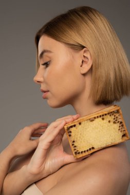 side view of blonde woman holding honeycomb in wooden frame isolated on grey  clipart