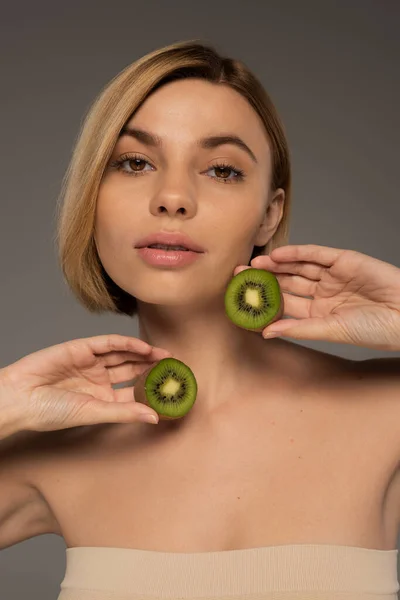 stock image young blonde woman with naked shoulders holding kiwi halves isolated on grey 