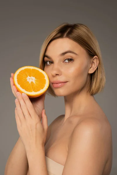 stock image young blonde woman with naked shoulders holding orange and looking at camera isolated on grey 