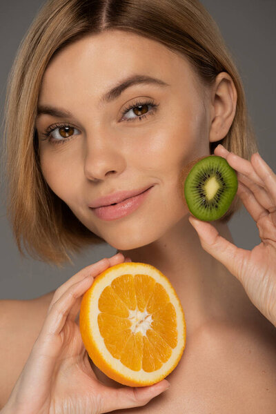 портрет веселой блондинки, держащей сочный апельсин и киви изолированные на сером 