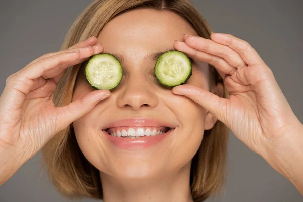 stock image portrait of cheerful woman covering eyes with sliced cucumbers isolated on grey 