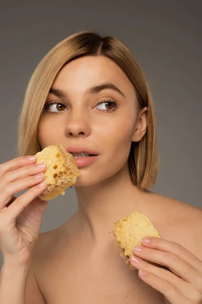 stock image young woman with bare shoulders holding pieces of sweet honeycomb while looking away isolated on grey