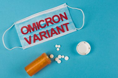 Top view of medical mask with omicron variant lettering near pills on blue background  clipart