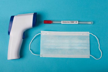 Top view of pyrometer near medical mask and cotton swab with omicron xbb lettering on blue background 