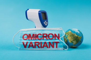 Pyrometer near medical mask with omicron variant lettering and globe on blue background 