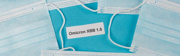 stock image Top view of omicron xbb lettering near protective masks on blue background, banner 