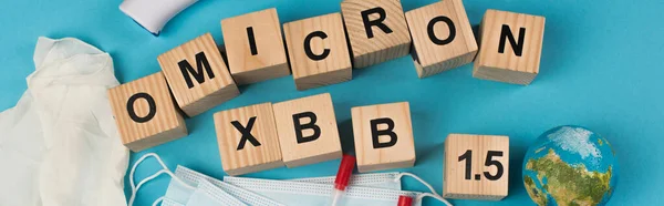 stock image Top view of wooden cubes with omicron xbb lettering near globe and medical masks on blue background, banner 