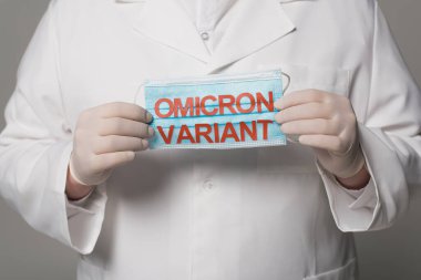 Cropped view of doctor in latex gloves holding medical mask with omicron variant lettering on grey background  clipart