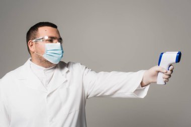 Doctor in protective goggles and mask holding pyrometer isolated on grey 