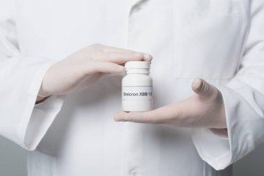 Cropped view of doctor in white coat and gloves holding jar with omicron xbb lettering isolated on grey 