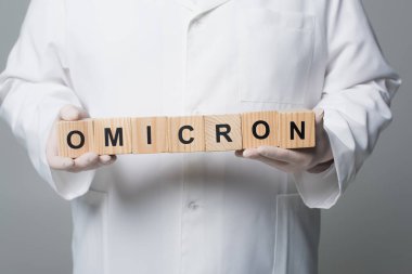 Cropped view of doctor in white coat and latex gloves holding wooden cubes with omicron lettering isolated on grey 